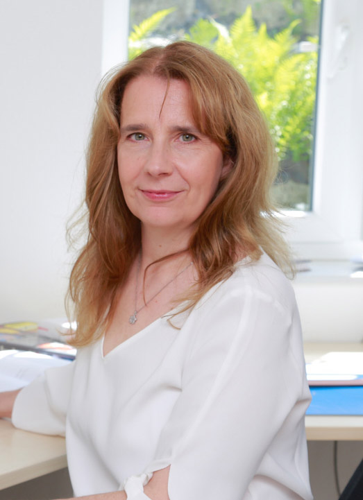 Website erstellen lassen. Silke Wengenroth-Lippke sitzt am Schreibtisch schaut direkt in die Kamera. Blonde mittellange Haare, grüne Augen freundliches Lächeln